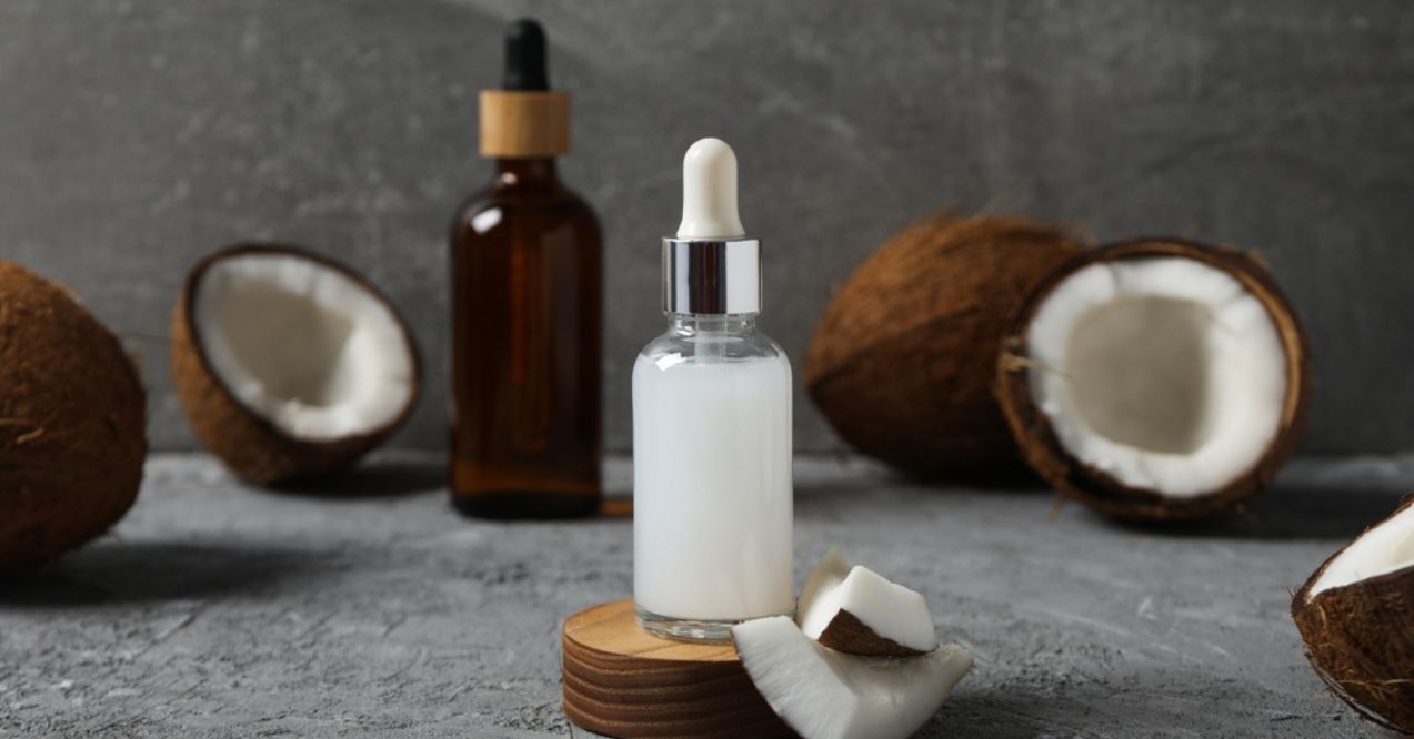 A dropper bottle of coconut oil placed in front of whole and halved coconuts