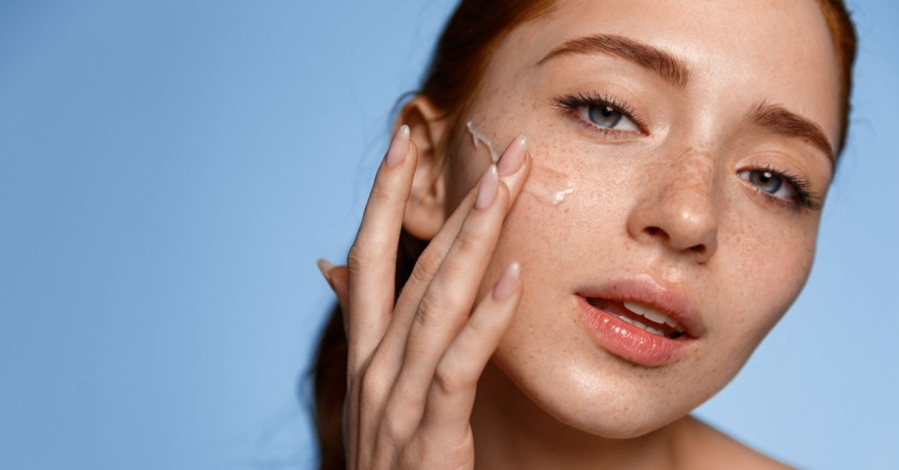 A young woman applying moisturizer to her face against a blue background