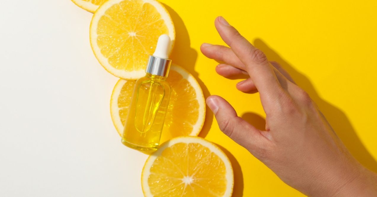 A hand reaching for a bottle of vitamin C serum placed on slices of fresh oranges