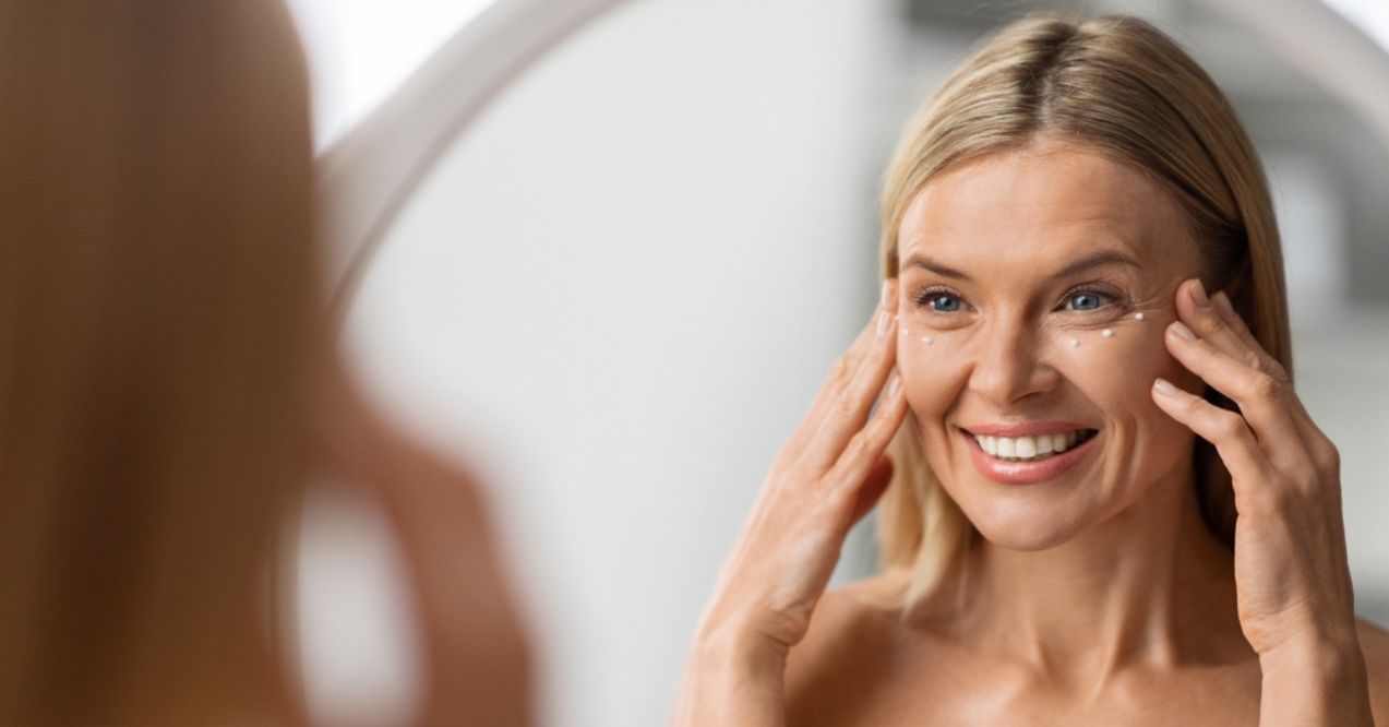 A smiling woman applying eye cream in front of a mirror