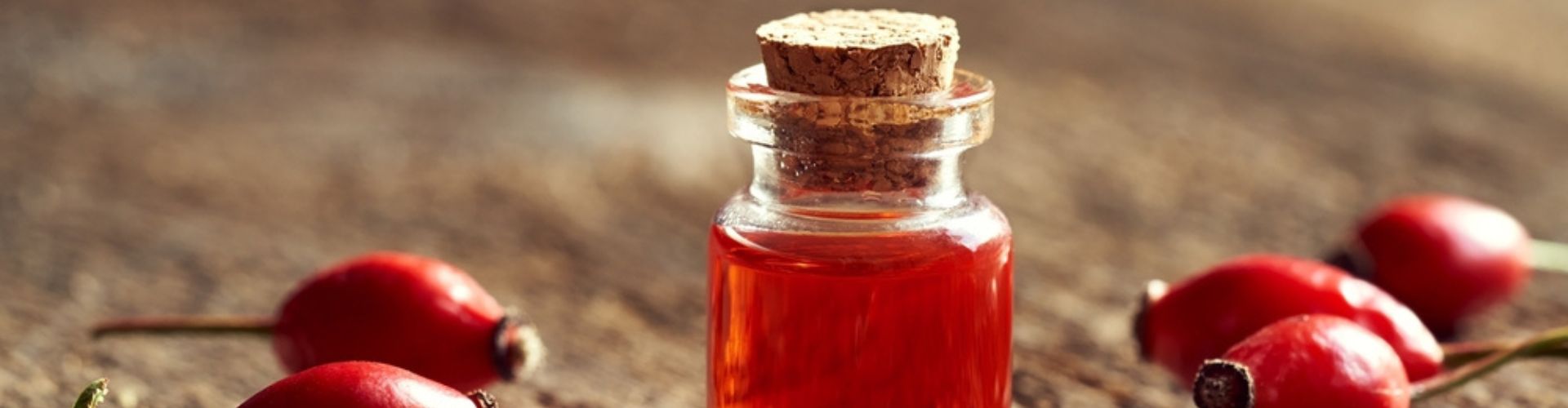 A small glass bottle of rosehip oil with a cork stopper, surrounded by fresh rosehips