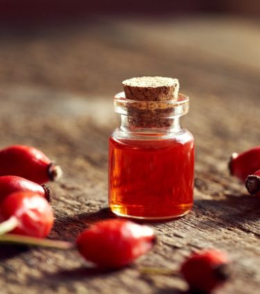 A small glass bottle of rosehip oil with a cork stopper, surrounded by fresh rosehips