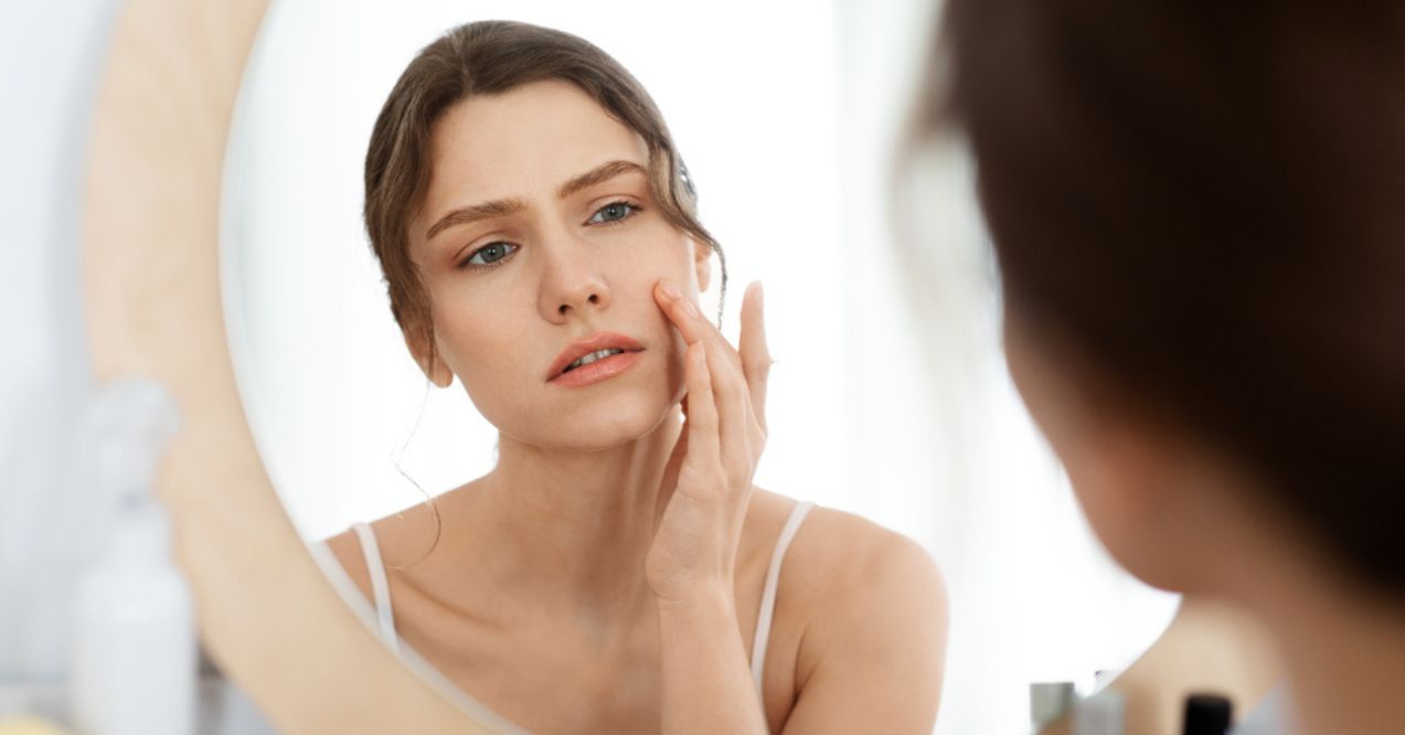 A young woman looking at her reflection in the mirror while touching her face with a concerned expression