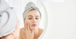 A woman with a towel wrapped around her head examining her face in the mirror