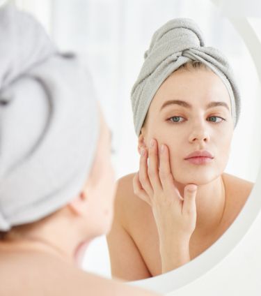 A woman with a towel wrapped around her head examining her face in the mirror