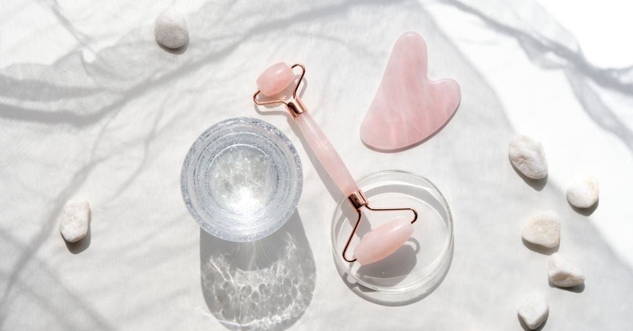 Rose quartz face roller and gua sha tool on a bright surface with small white stones