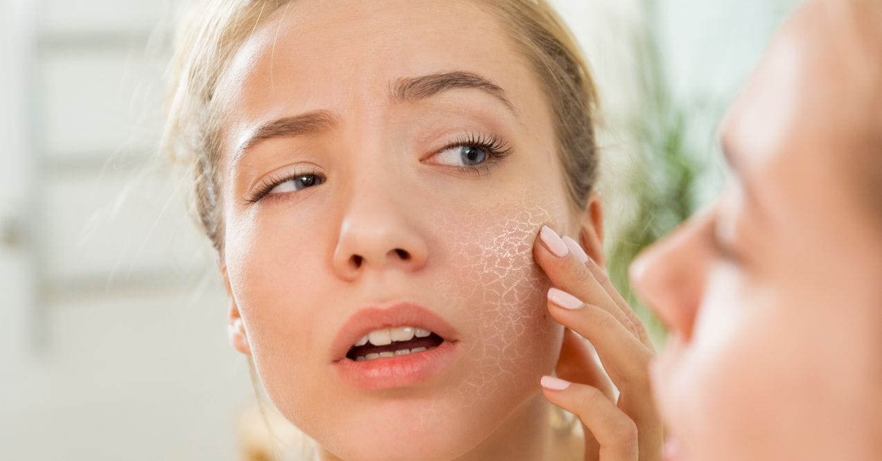 Woman examining dry, cracked skin on her cheek in the mirror.