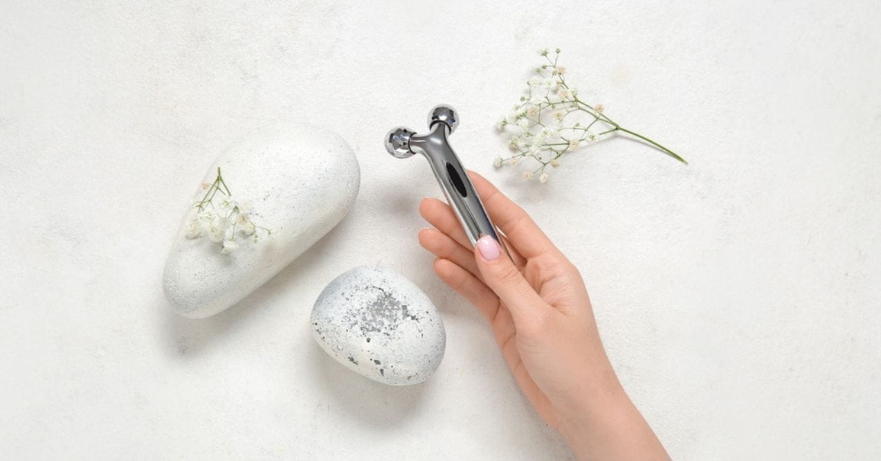 Hand holding a metal face roller beside smooth stones and flowers