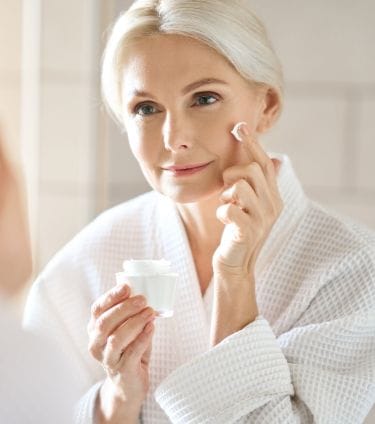 Smiling mature woman in a white robe applying face cream in front of a bathroom mirror.