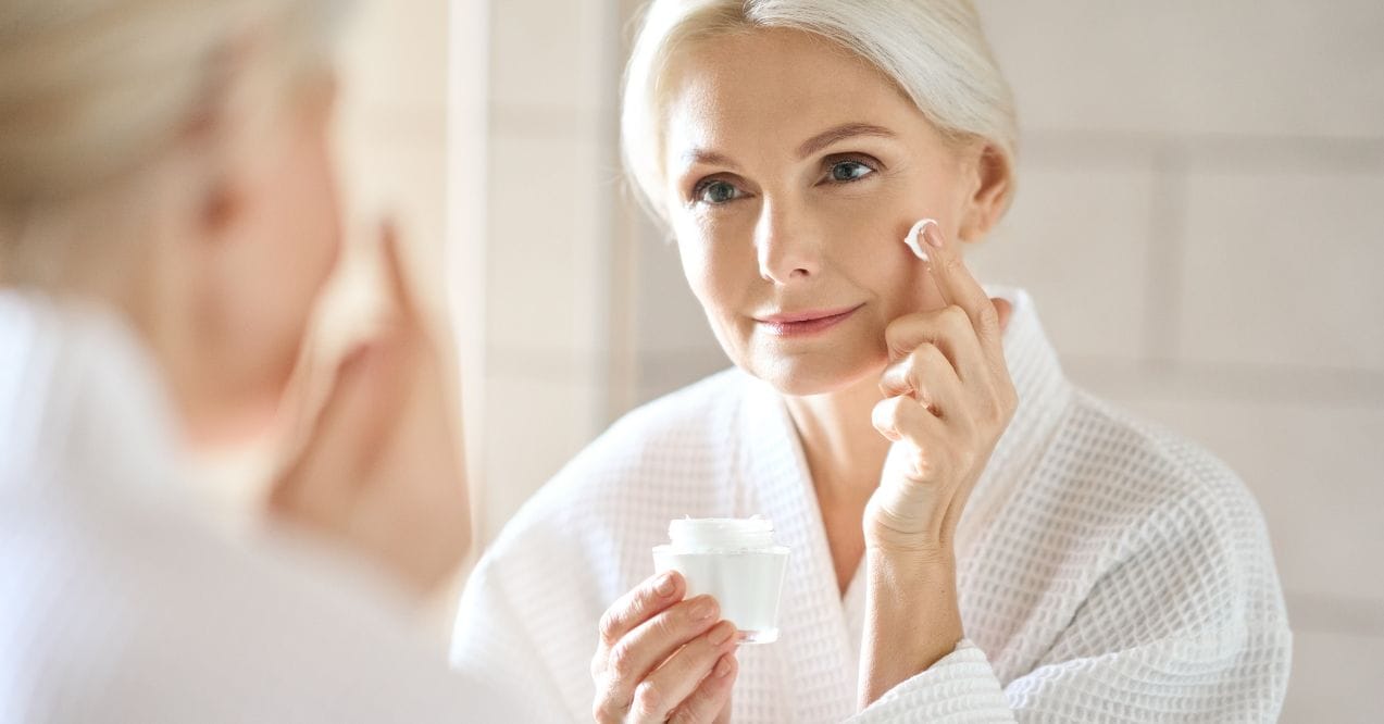 Smiling mature woman in a white robe applying face cream in front of a bathroom mirror.