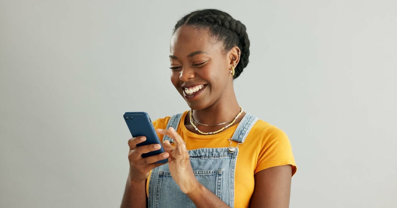 Smiling woman using a smartphone, wearing a yellow top and denim overalls.