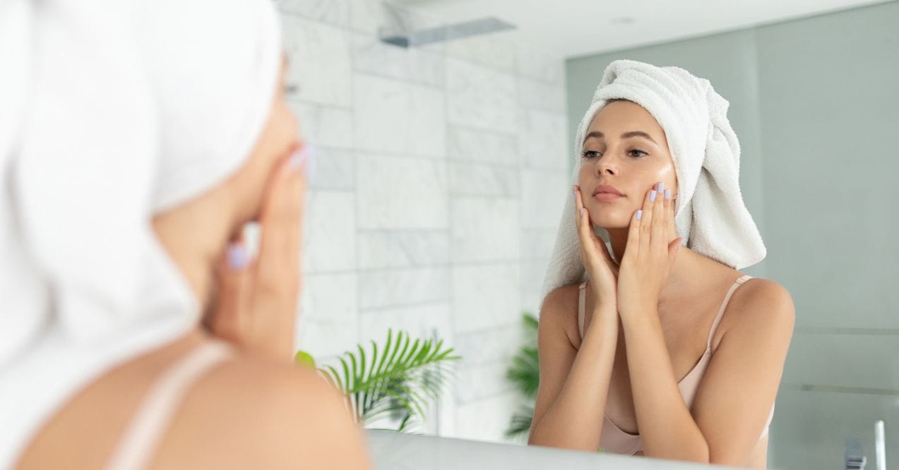 Woman with a towel on her head gently applying moisturizer while looking in the mirror.