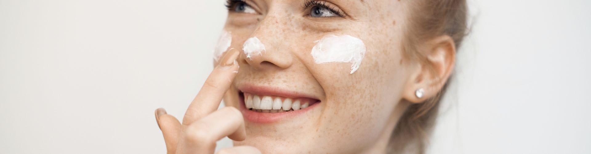 Smiling woman applying moisturizer to her face with visible freckles.