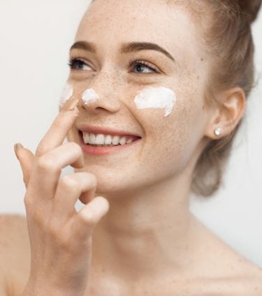 Smiling woman applying moisturizer to her face with visible freckles.