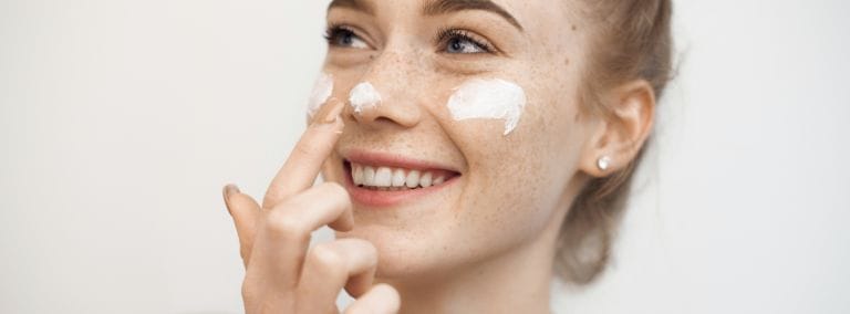 Smiling woman applying moisturizer to her face with visible freckles.