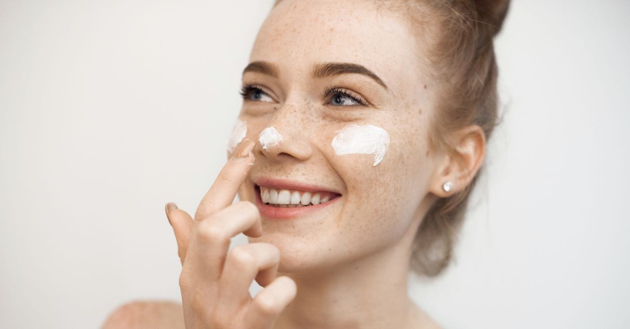 Smiling woman applying moisturizer to her face with visible freckles.