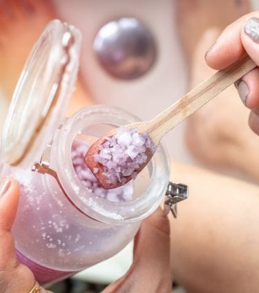 A close-up of purple bath salt being scooped, answering the question, is salt water good for your skin.