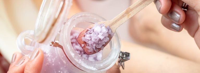 A close-up of purple bath salt being scooped, answering the question, is salt water good for your skin.