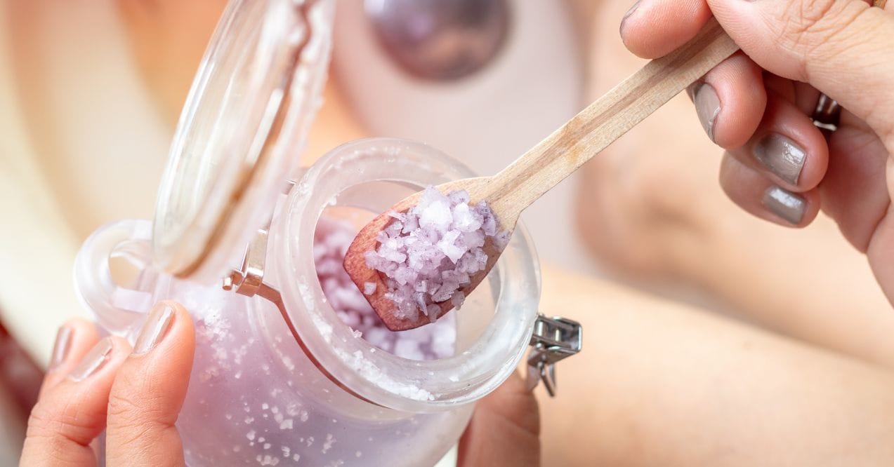 A close-up of purple bath salt being scooped, answering the question, is salt water good for your skin.