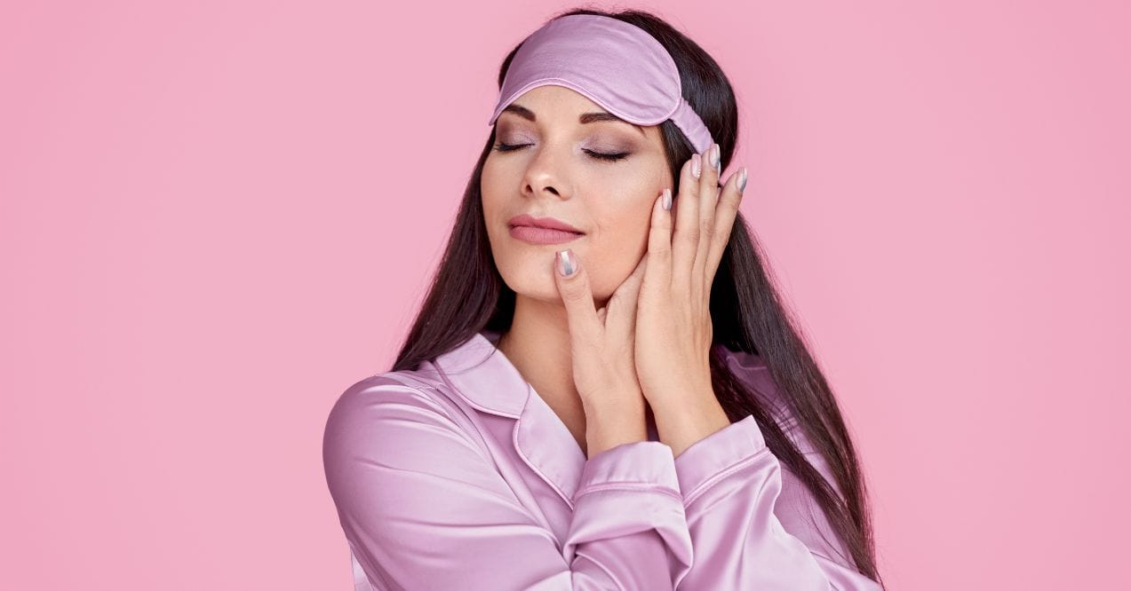 Relaxed woman in pink pajamas and a sleep mask caressing her face before bedtime.