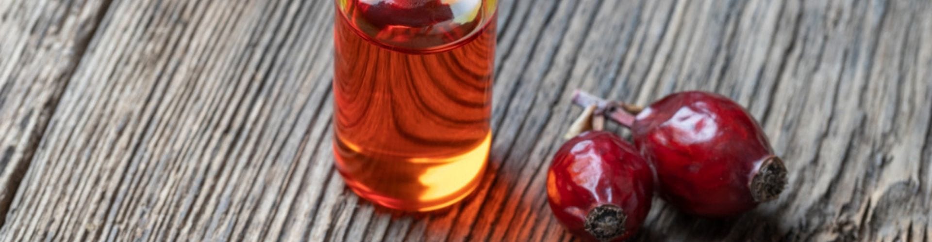 Small glass bottle of rosehip oil with fresh rosehips on a rustic wooden surface