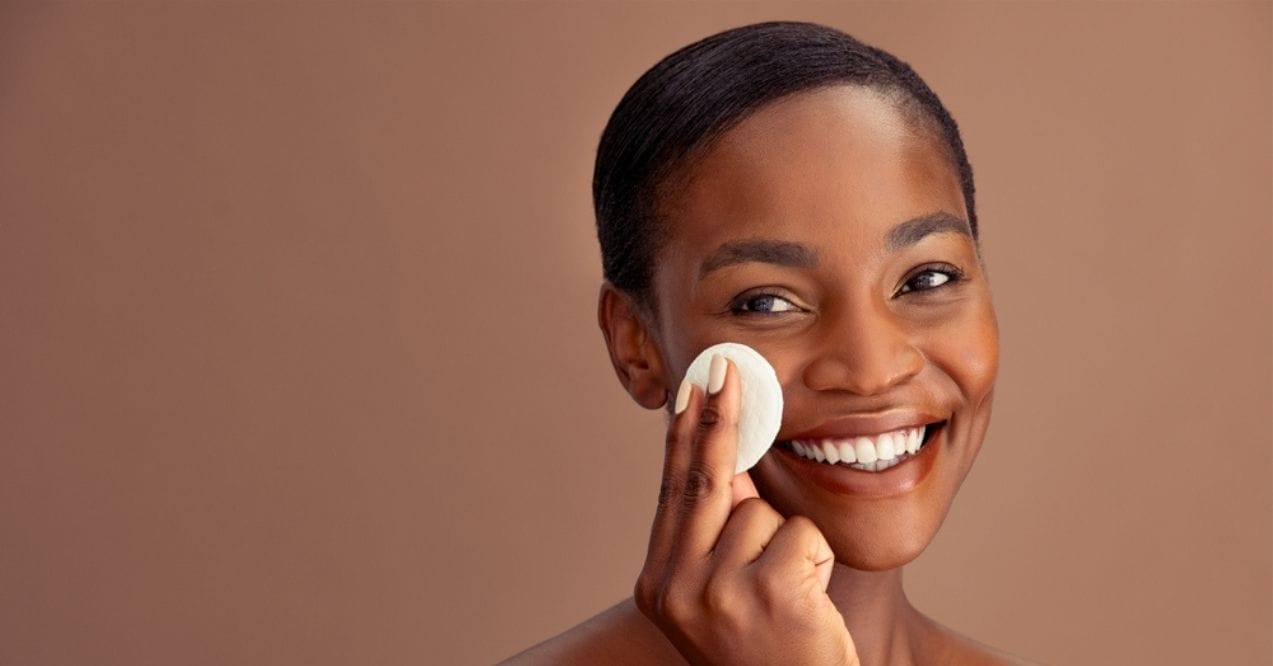 Smiling woman using a cotton pad to cleanse her face against a brown background