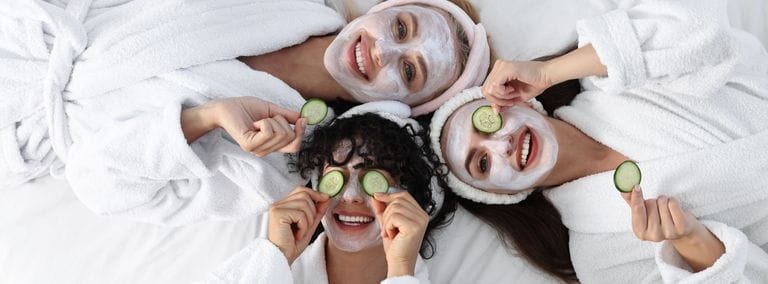 Three women in spa robes with face masks and cucumber slices