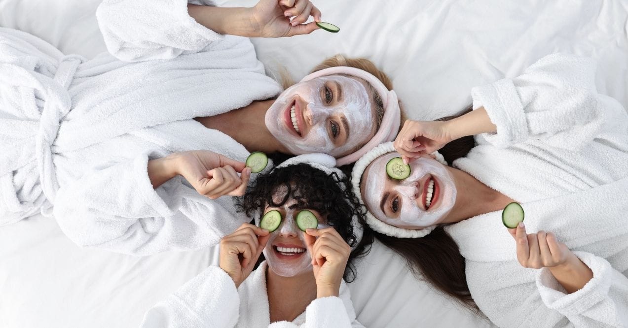 Three women in spa robes with face masks and cucumber slices